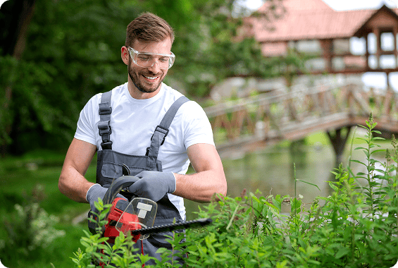 Man wearing a t-shirt, black gloves, and glasses, using a hedge trimmer to trim bushes in front of a bridge